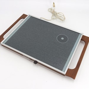 May include: A vintage electric griddle with a dark gray cooking surface and a silver frame. The griddle has wooden handles on each side and a control knob. A power cord with a plug is connected to the griddle.