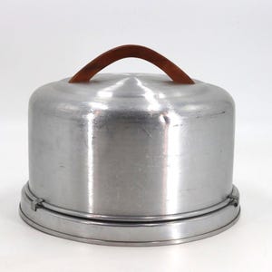 May include: A round, brushed aluminum cake carrier with a smooth, domed lid and a flat base. The lid features a curved, brown wooden handle attached to the top center. The two pieces are secured together by small metal clips along the bottom edge.