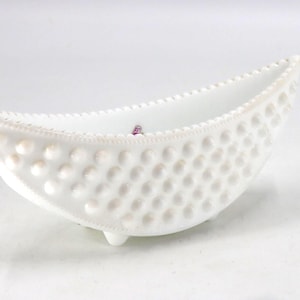 May include: A white, oval-shaped dish with a textured, hobnail pattern. The dish has a scalloped rim and rests on three small feet. The interior is smooth and empty, with a small object inside.