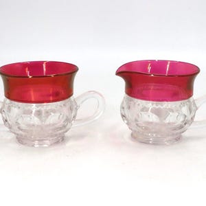 May include: Two vintage glass cream and sugar set. The clear glass bases have a diamond pattern, and the rims are a vibrant red. Each piece has a clear handle.
