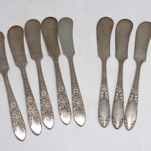 May include: A set of eight silver-toned metal butter knives arranged in two groups. Each knife features a wide, rounded blade and a handle decorated with an embossed floral pattern that extends from the base of the blade to the tip of the handle.