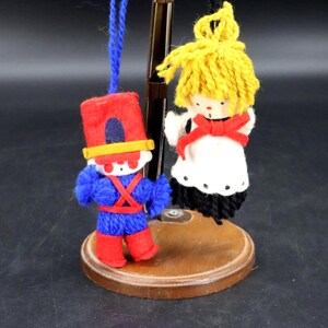 May include: Two handmade yarn dolls on a wooden stand. One doll is dressed as a soldier in red and blue, the other as an angel in white and black. The soldier doll has a red hat and the angel doll has yellow yarn hair.