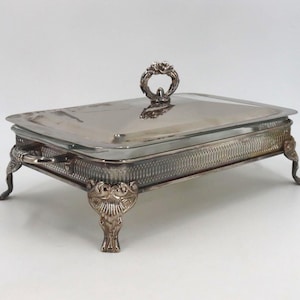 May include: A rectangular silver-plated serving dish with a removable lid featuring a decorative handle. The base has a textured lattice pattern along the sides, two side handles, and four ornate feet with detailed scrollwork and leaf motifs.