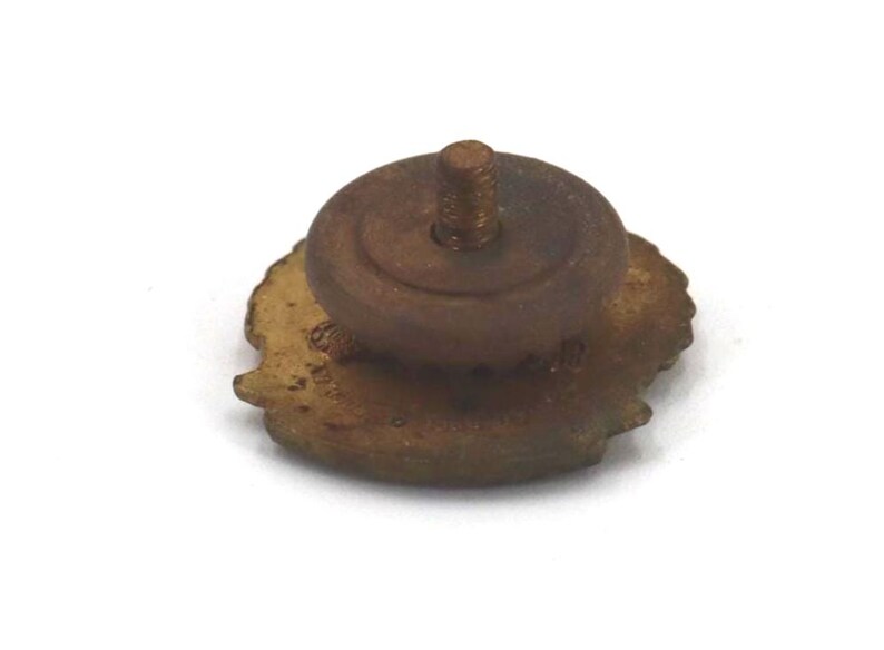May include: A vintage-style, bronze-colored pin with a threaded post and a circular base. The pin has a detailed, possibly floral, design around the edge. The pin is set against a white background.