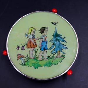 May include: A round, vintage trinket box with a light green lid featuring a painted scene of two children and a tree. The children are walking on grass, and a bird is perched atop the tree. The box has a silver rim and red accents.