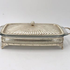May include: Silver-plated serving dish with a rectangular shape and a lid. The lid has a sunburst design and a small round handle. The dish has handles on each side and sits on decorative feet.