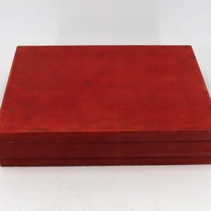 May include: A rectangular wooden box with a flat lid and a deep red finish. The box features a smooth, solid surface with visible wood grain texture and a horizontal seam where the lid meets the base, creating a simple, clean, and functional design.