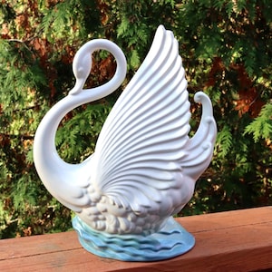 May include: A white ceramic swan figurine with a graceful, curved neck and detailed wing feathers. The swan is perched on a light blue base, creating a decorative accent piece. The figurine is set against a backdrop of green foliage.