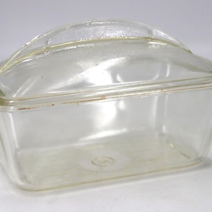 May include: A clear, rectangular glass loaf dish with a matching lid. The lid has a curved handle. The dish is empty and sits on a white surface. The glass has a slight yellow tint.