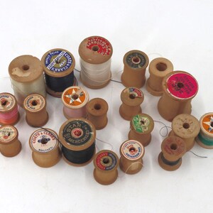 May include: A collection of vintage wooden spools of thread in various sizes and colors. Some spools have labels with text, and the threads are in shades of white, pink, black, and green. The spools are arranged on a white surface.