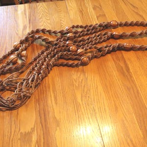 May include: A brown macrame plant hanger constructed from twisted cord featuring integrated wooden beads throughout the length of the strands. The hanger terminates in a long, loose fringe tassel at the bottom and includes a circular ring at the top.