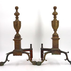 May include: Pair of antique brass and bronze andirons. Each andiron features a decorative urn-shaped top, a square base, and curved legs with a claw foot design. The andirons are designed to hold logs in a fireplace.