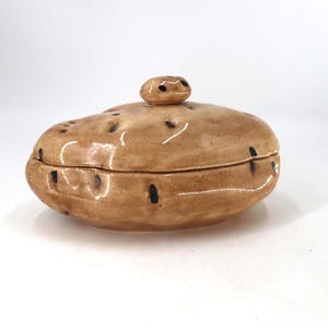 May include: A ceramic lidded container shaped like a potato, featuring a glossy tan finish with dark brown spots and an oval knob on top. The lidded dish has an irregular, organic form with a smooth, reflective surface and a distinct horizontal seam.