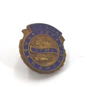 May include: Vintage brass-toned pin with blue lettering and accents. The pin features the text "DIVISION 1287" and "A.A.S.E.R. & M.C.E. OF A.F. OF L." A detailed illustration of a vehicle is in the center of the pin.