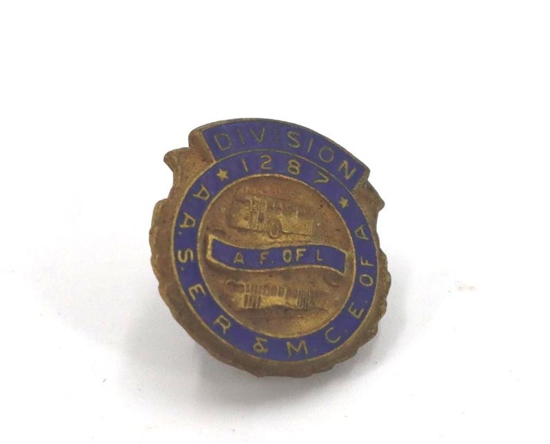 May include: Vintage brass-toned pin with blue lettering and accents. The pin features the text "DIVISION 1287" and "A.A.S.E.R. & M.C.E. OF A.F. OF L." A detailed illustration of a vehicle is in the center of the pin.