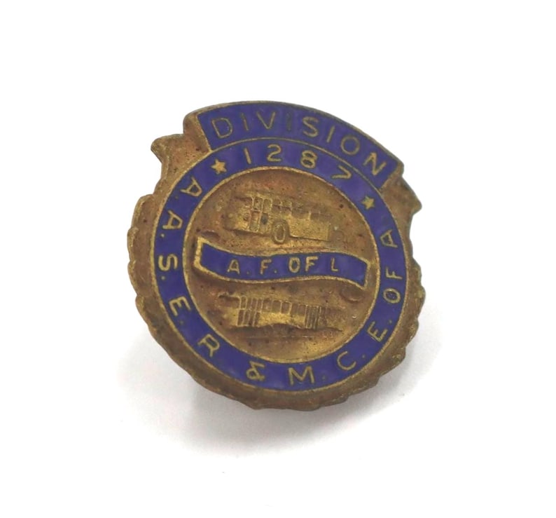 May include: A vintage brass-toned pin with blue enamel lettering and accents. The pin features the text "DIVISION 1287" and "A.A.S.E.R. & M.C.E. A.F. OF L." in a circular design with a central emblem.