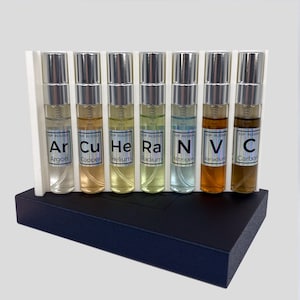 May include: A set of seven glass vials with silver caps, each containing a different colored liquid. The vials are labeled with chemical symbols and names: Argon, Copper, Helium, Radium, Nitrogen, Vanadium, and Carbon. The vials are displayed in a white and black display case.