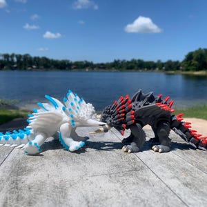 May include: Two articulated dragon figurines facing each other. One is white with bright blue spikes and accents, while the other is dark gray with vibrant red spikes and accents. Both creatures feature detailed scales, horns, and tails.