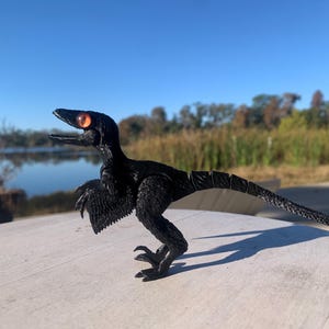 Puede incluir: Figura de dinosaurio negro con ojos naranjas, posada sobre una superficie clara. El juguete tiene un acabado texturizado y la boca abierta. El fondo muestra un lago y árboles bajo un cielo azul.