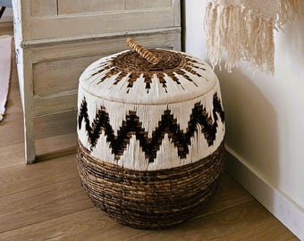 May include: A round storage basket with a woven design. The basket features a natural brown base, a white and brown patterned middle section, and a woven lid with a handle. The basket is made of natural materials and is suitable for home decor.