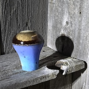 May include: A handcrafted ceramic bottle stopper with a gradient blue base, transitioning to brown and gold at the top. The stopper is displayed on a weathered wooden surface, showcasing its unique design and texture.