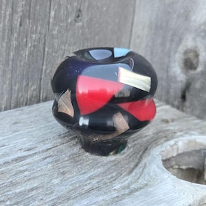 May include: A decorative, handcrafted glass knob with a black base, red and gold accents, and a glossy finish. The knob has a unique, rounded shape and is suitable for furniture or cabinet doors.