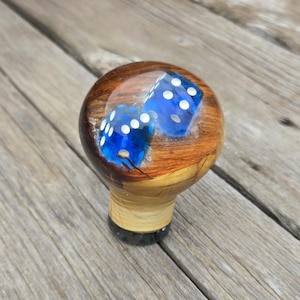 May include: A wooden shift knob with two blue dice encased in clear resin. The dice have white dots. The knob has a light brown wooden base and a dark brown top. The background is a wooden surface.