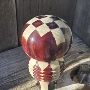 May include: A decorative wooden object with a spherical top and a cylindrical base. The top features a checkerboard pattern in red and natural wood tones. The base has a ribbed design. The object is displayed on a wooden surface.