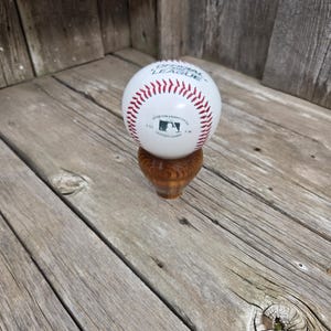 May include: A white baseball with red stitching, labeled "OFFICIAL LEAGUE" and "LEATHER COVER", sits atop a turned wooden base. The baseball has the MLB logo. The background is weathered wood.