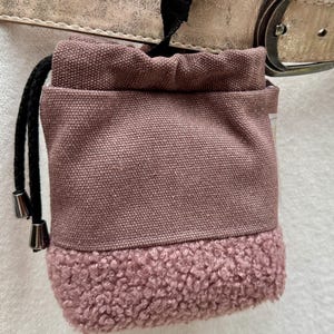 Puede incluir: Una bolsa de golosinas para perros de color malva con cierre de cordón, sujeta a un collar dorado para perros. La parte inferior de la bolsa tiene un material texturizado y difuso. Una correa y hebilla negras aseguran la bolsa al collar.