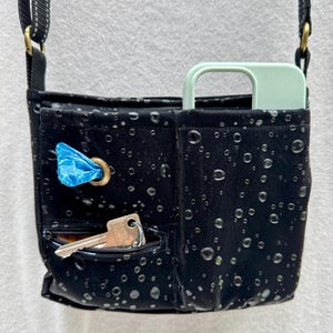 Puede incluir: Bolso bandolera negro con un estampado de gotas de agua. Cuenta con un bolsillo frontal con cremallera con una llave, un dispensador de bolsas de residuos azul y un bolsillo que contiene un teléfono verde claro. El bolso tiene una correa negra con herrajes dorados.