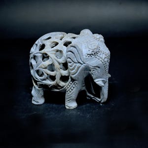 Puede incluir: Una escultura de elefante de piedra gris tallada con intrincados patrones calados en su cuerpo. El elefante tiene tallas detalladas en la cabeza y la trompa, y se apoya sobre cuatro patas. El fondo es oscuro, resaltando los detalles de la escultura.