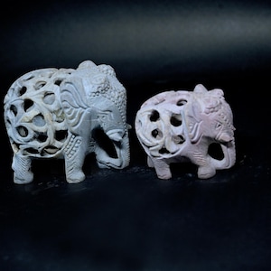 Puede incluir: Dos figuras de elefantes de piedra tallada. El elefante más grande es de color gris claro y el más pequeño es rosa. Ambos tienen intrincados diseños calados y están sobre un fondo oscuro. Los elefantes tienen colmillos tallados y adornos decorativos en la cabeza.