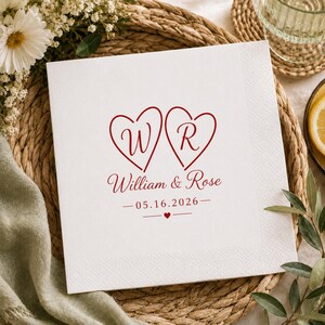May include: A square white paper napkin featuring a red printed design of two hearts containing the letters W and R. Below the hearts, the text reads William & Rose, followed by the date 05.16.2026 and a small red heart centered between two lines.