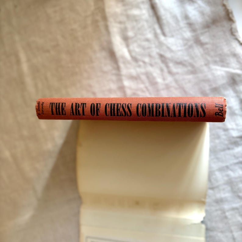 May include: A vintage book titled "THE ART OF CHESS COMBINATIONS" with an orange cover and black lettering. The book is lying on top of a stack of white pages, with a neutral-colored fabric in the background.