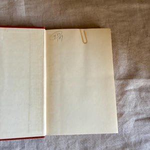 May include: Open book with red cover and cream-colored pages. A gold-colored paper clip secures a page. Handwritten numbers and symbols are visible on the upper left page. The book is open on a neutral-colored surface.