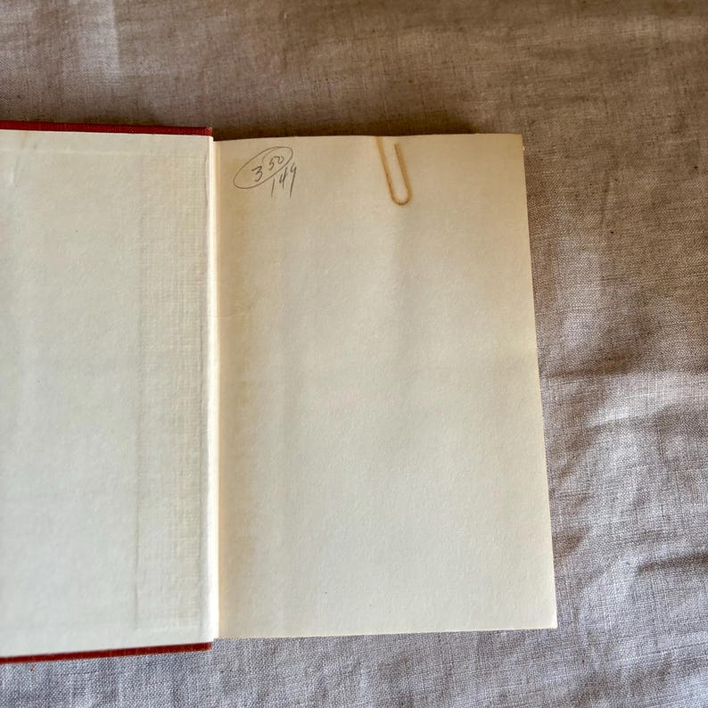 May include: Open book with red cover and cream-colored pages. A gold-colored paper clip secures a page. Handwritten numbers and symbols are visible on the upper left page. The book is open on a neutral-colored surface.