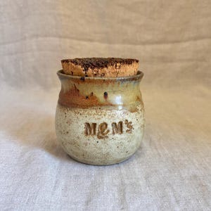 May include: A small, handcrafted ceramic jar with a cork stopper. The jar is beige with brown and green accents and the word "MOM'S" is embossed on the front. The cork stopper is dark brown.