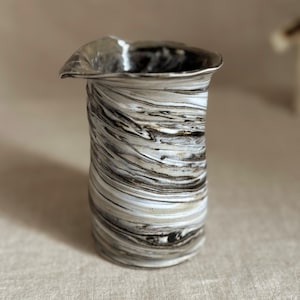 May include: A ceramic vase with a unique, swirled design in shades of black, white, and gray. The vase has a flared, asymmetrical rim and a cylindrical body. The textured surface gives it a handcrafted appearance.