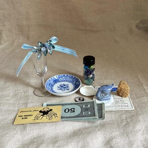 May include: A collection of decorative items including a champagne flute with a blue and crystal embellishment, a blue and white patterned dish, a small bottle filled with beads, and Monopoly money. Also included are a seashell, a small ceramic whale, and a rock.