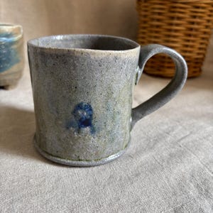 May include: A handmade ceramic mug with a speckled, gray-blue glaze. The mug features a rounded handle and a small, dark blue floral design. The mug is part of a pottery collection and is suitable for hot beverages.