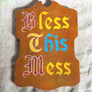 May include: Wooden sign with the words "Bless This Mess" in a decorative font. The sign is brown with the words in red, yellow, and blue. The sign has a unique shape with a curved top.