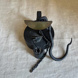 May include: Antique black metal oil lamp with a brass bowl-shaped reservoir. The lamp features a chain, a curved hook, and a handle. The design is ornate and vintage, suggesting a historical or decorative item.