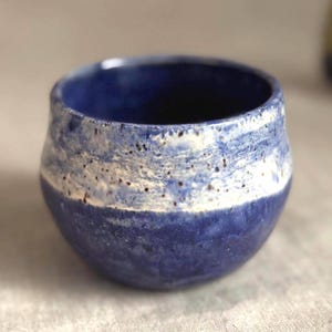 May include: A handmade ceramic cup with a rounded shape. The cup features a deep blue base, a white band with speckled details, and a darker blue rim. The interior is a deep blue color.