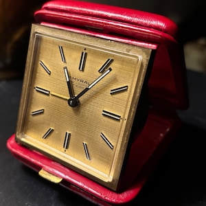 May include: A vintage gold-toned travel clock with a square face and black hands and markers. The clock is encased in a red leather folding case. The clock face has the text "AMYRAL" and "7 RUBIS".