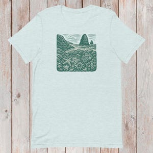 Peut inclure: T-shirt bleu clair avec un motif vert représentant une scène côtière. Le motif comprend une plage, l'océan, des rochers et une étoile de mer. Le t-shirt est en matière douce et a des manches courtes.