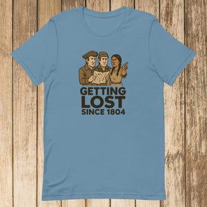May include: Light blue t-shirt featuring a graphic of three people looking at a map, with the text "Getting Lost Since 1804." The illustration is in shades of brown and tan. The shirt is made of a soft material.