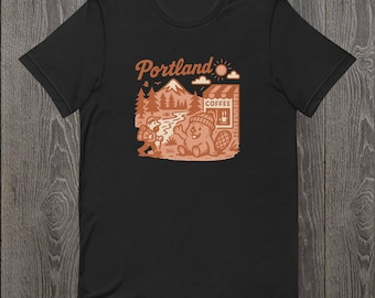 Könnte beinhalten: Schwarzes T-Shirt mit einem Grafikdesign, das das Wort "Portland" über einer Szene mit einem Berg, Bäumen, einem Biber und einem Café zeigt. Der Biber trägt einen Hut und winkt. Eine Person wandert in der Nähe.