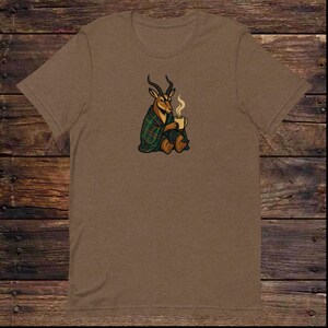 Könnte beinhalten: Braunes T-Shirt mit einer Illustration eines Antilopen, die in eine grün-schwarz karierte Decke gehüllt ist und eine dampfende Tasse hält. Die Antilope sitzt. Das T-Shirt hat einen Rundhalsausschnitt.