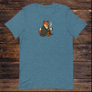 Könnte beinhalten: Blaues T-Shirt mit einem Cartoon-Fuchs-Design. Der Fuchs ist in eine grün, blau und braun karierte Decke gehüllt und hält eine dampfende Tasse. Der Fuchs ist orange und weiß. Das T-Shirt ist aus weichem Material.
